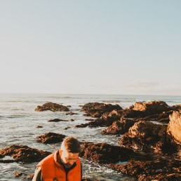 man wearing orange and black bubble jacket standing on rock while looking down viewing calm body of water during daytime